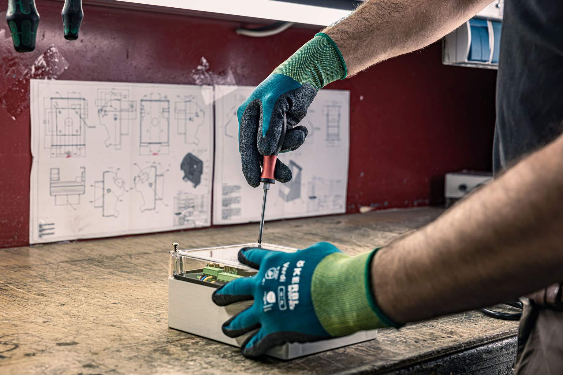 Un employé travaille sur un établi à l'aide d'un tournevis sur un composant électronique, tout en portant des gants de travail verts-bleus